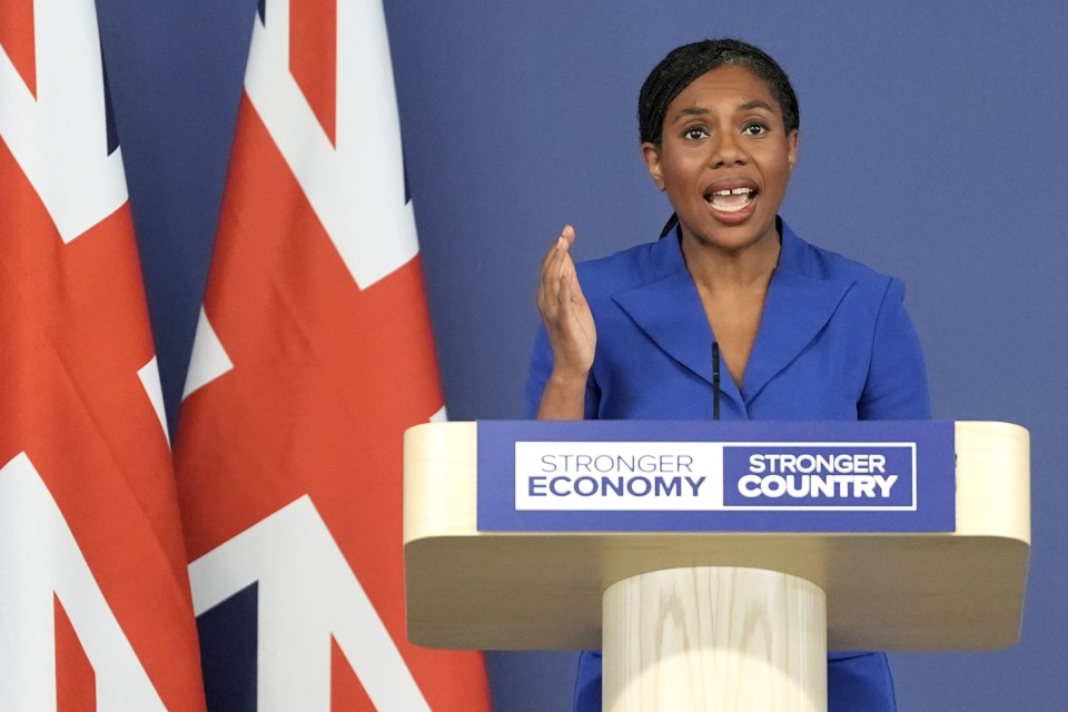 Conservative Party Leader Kemi Badenoch speaking at a podium with British flags in the background.