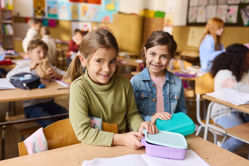 Two elementary school girls opening their lunch boxes in a classroom.