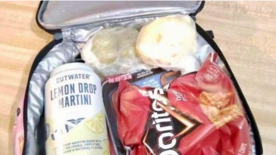 An open lunchbox contains a can of "Cutwater Lemon Drop Martini," two wrapped food items, and a bag of Doritos.