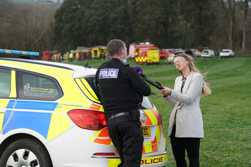 A police officer is interviewed by a journalist next to a police car in a grassy field.
