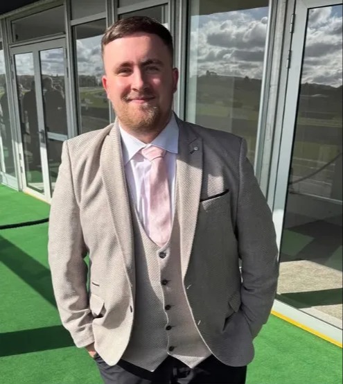 Luke Littler wearing a light-colored suit with a pink tie and vest at Cheltenham Racecourse.