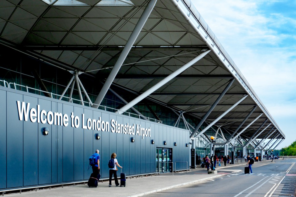 Passengers with luggage walking towards the entrance of London Stansted Airport with a welcome sign.