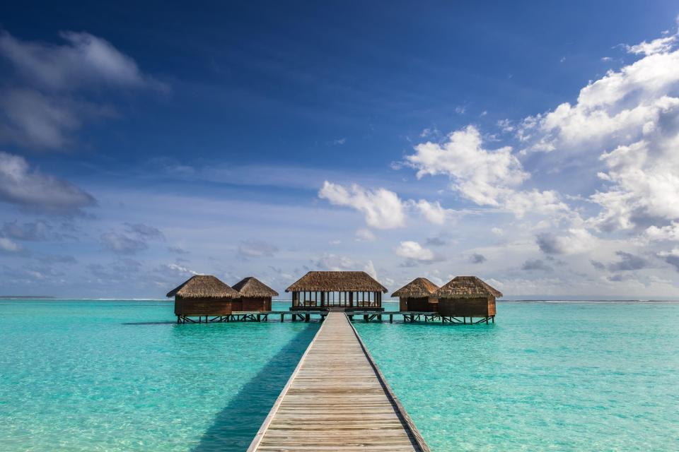 A wooden boardwalk leading to several thatched-roof bungalows over turquoise water in the Maldives.