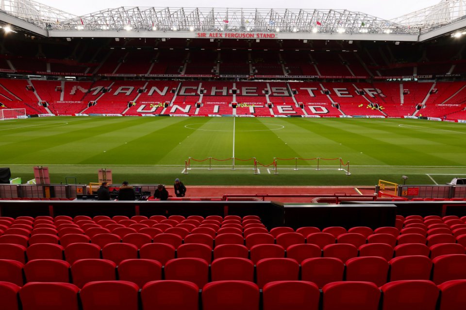 The Manchester United football stadium, with rows of empty red seats in the foreground and the green pitch beyond.