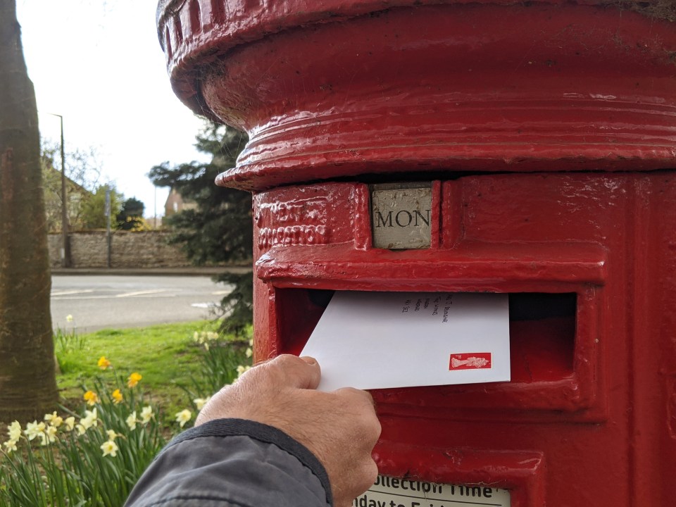 Hand posting a stamped letter into a red post box in a rural setting.