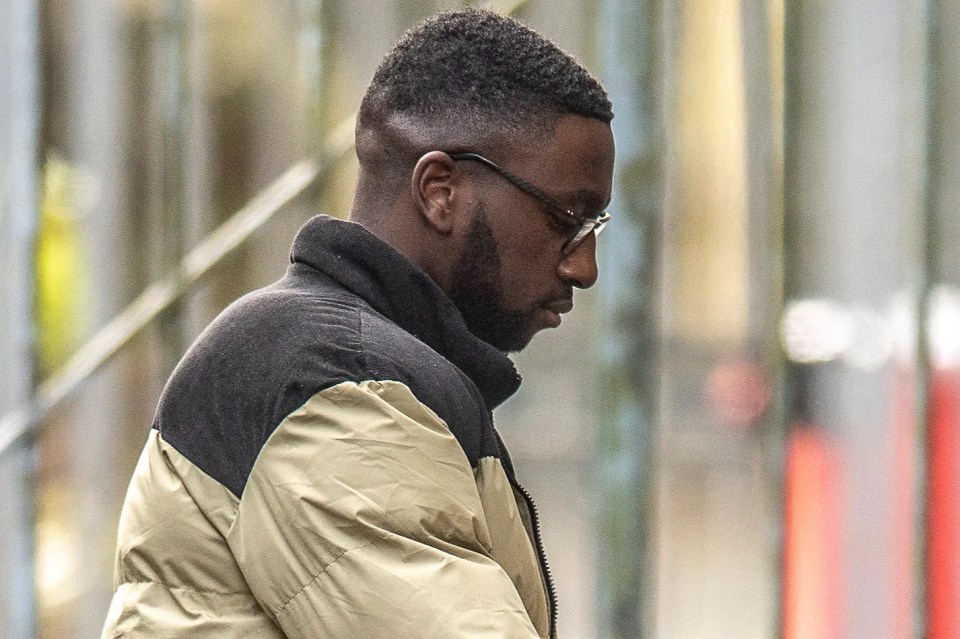 Gift Oladele in a black and tan jacket and glasses, looking downcast.