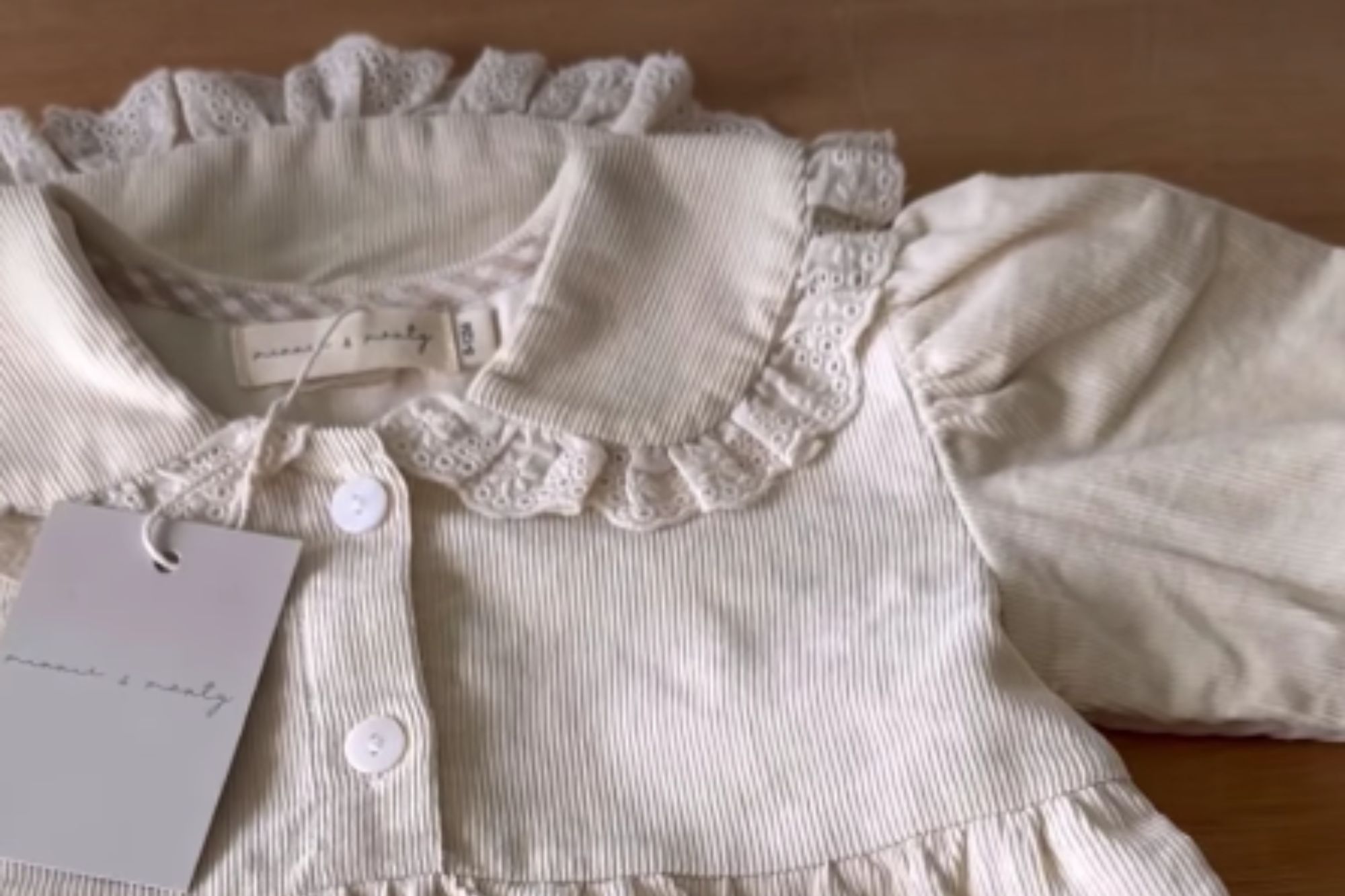 An image collage containing 1 images, Image 1 shows A light beige baby romper with a ruffled collar, long sleeves, and snaps at the crotch