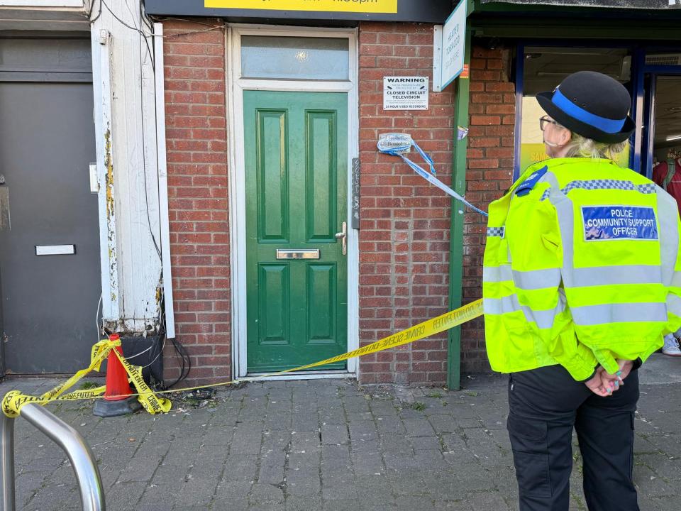 A police community support officer stands in front of an address with crime scene tape.