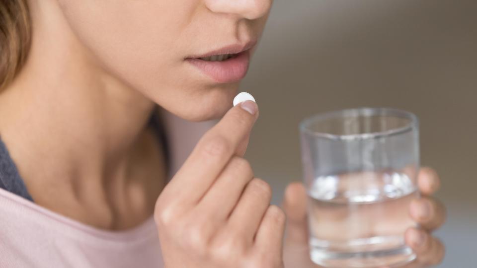Close up of a woman about to take a pill, holding it to her mouth with one hand, and holding a glass of water in the other.