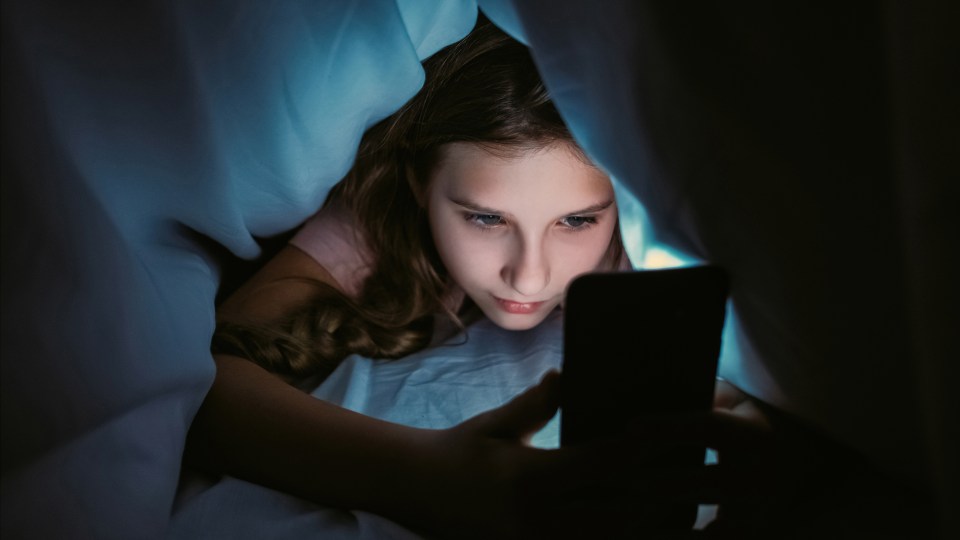 A young girl under bedcovers uses a smartphone, its light illuminating her face.