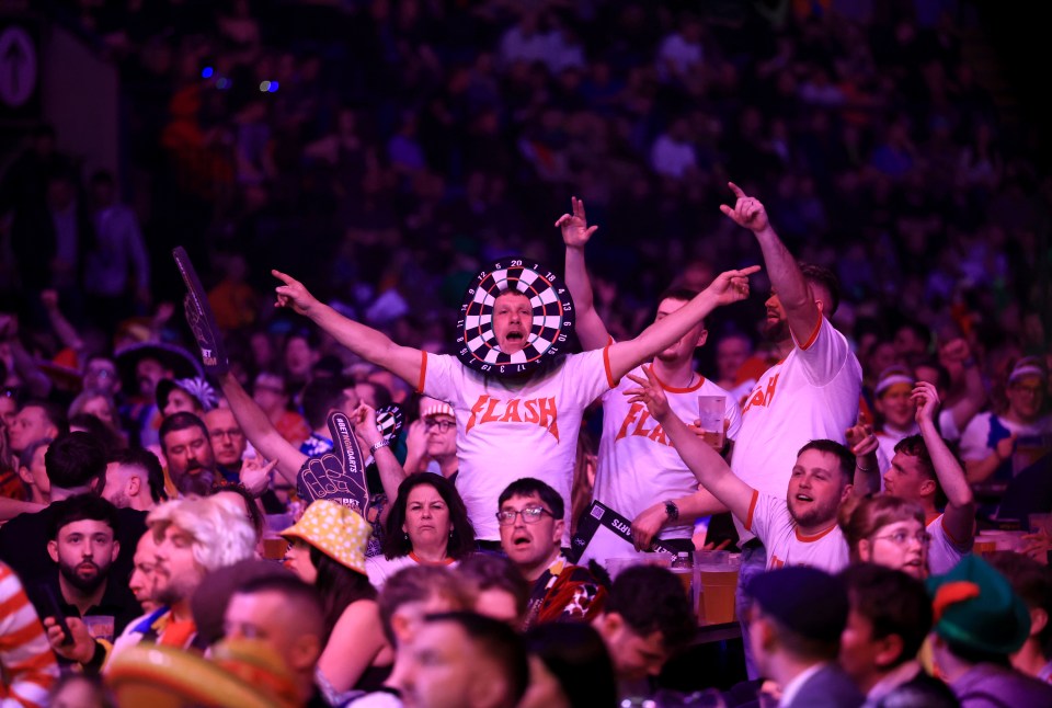 A man in the crowd at the BetMGM Premier League darts event wearing a dartboard around his face and a "Flash" shirt, with his arms outstretched.