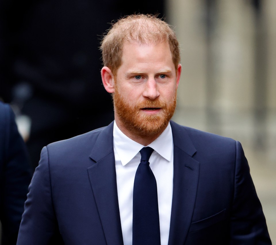 Prince Harry in a navy suit with a white shirt and dark blue tie, looking forward with a serious expression.