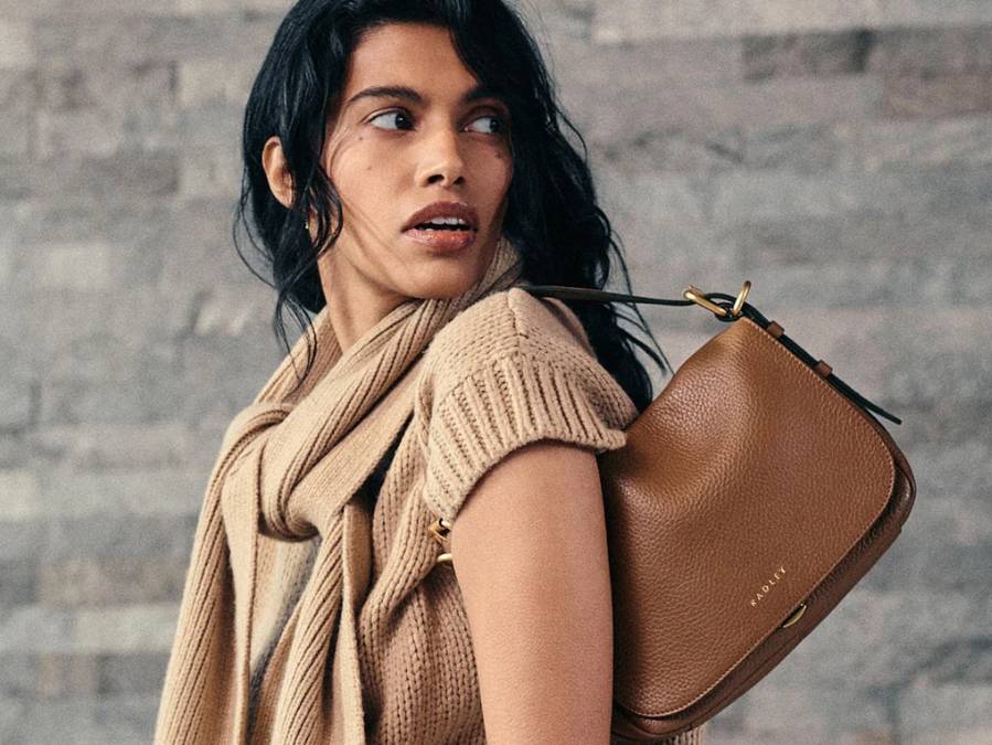 A woman wearing a tan knit sweater and scarf, holding a tan Radley shoulder bag.
