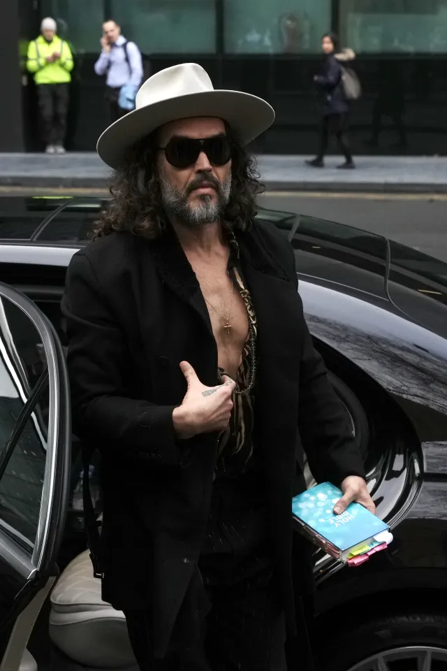 Russell Brand in a hat and sunglasses, wearing an unbuttoned shirt and black coat, holding a Bible, outside Southwark Crown Court.