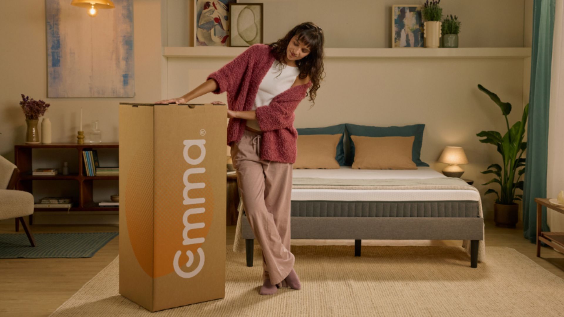 A woman standing next to a tall Emma mattress box in a bedroom.