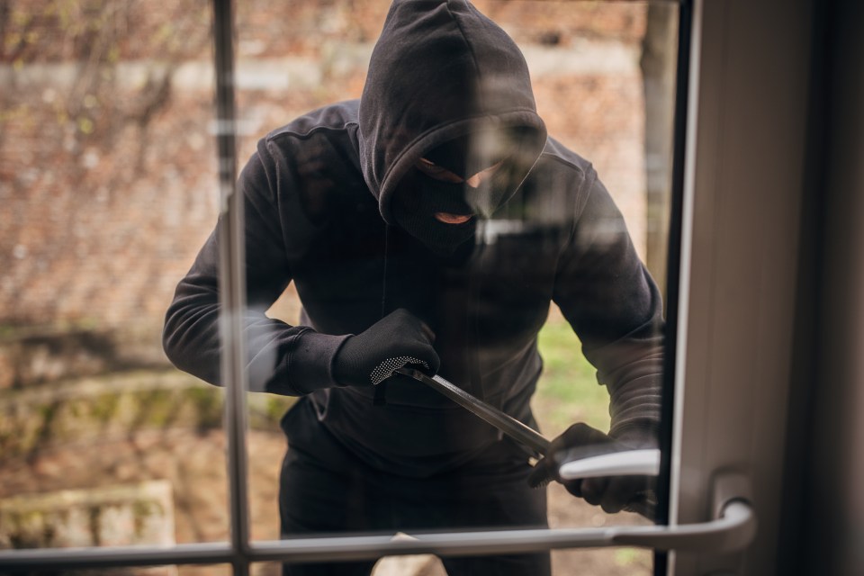 A masked burglar in a black hoodie and gloves uses a crowbar to pry open a house door.