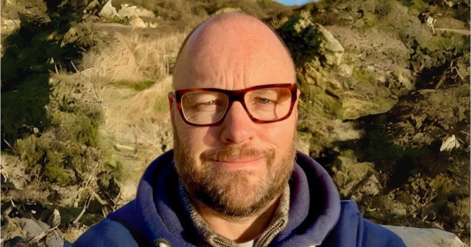 Dr. Simon Newman smiling in front of a hilly, vegetated background.
