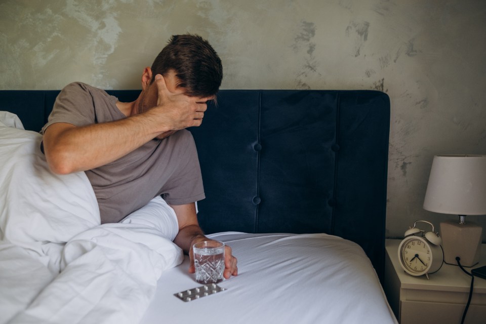Man in bed with a headache covering his eyes, reaching for water and pills.