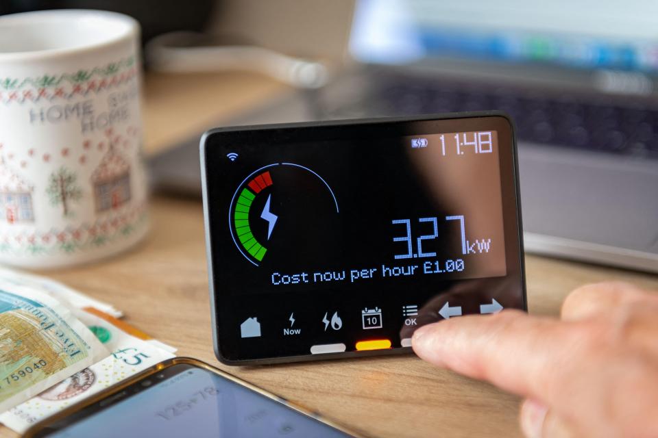 A person checking a smart meter displaying a rising energy cost of £1.00 per hour, with money and a calculator nearby.