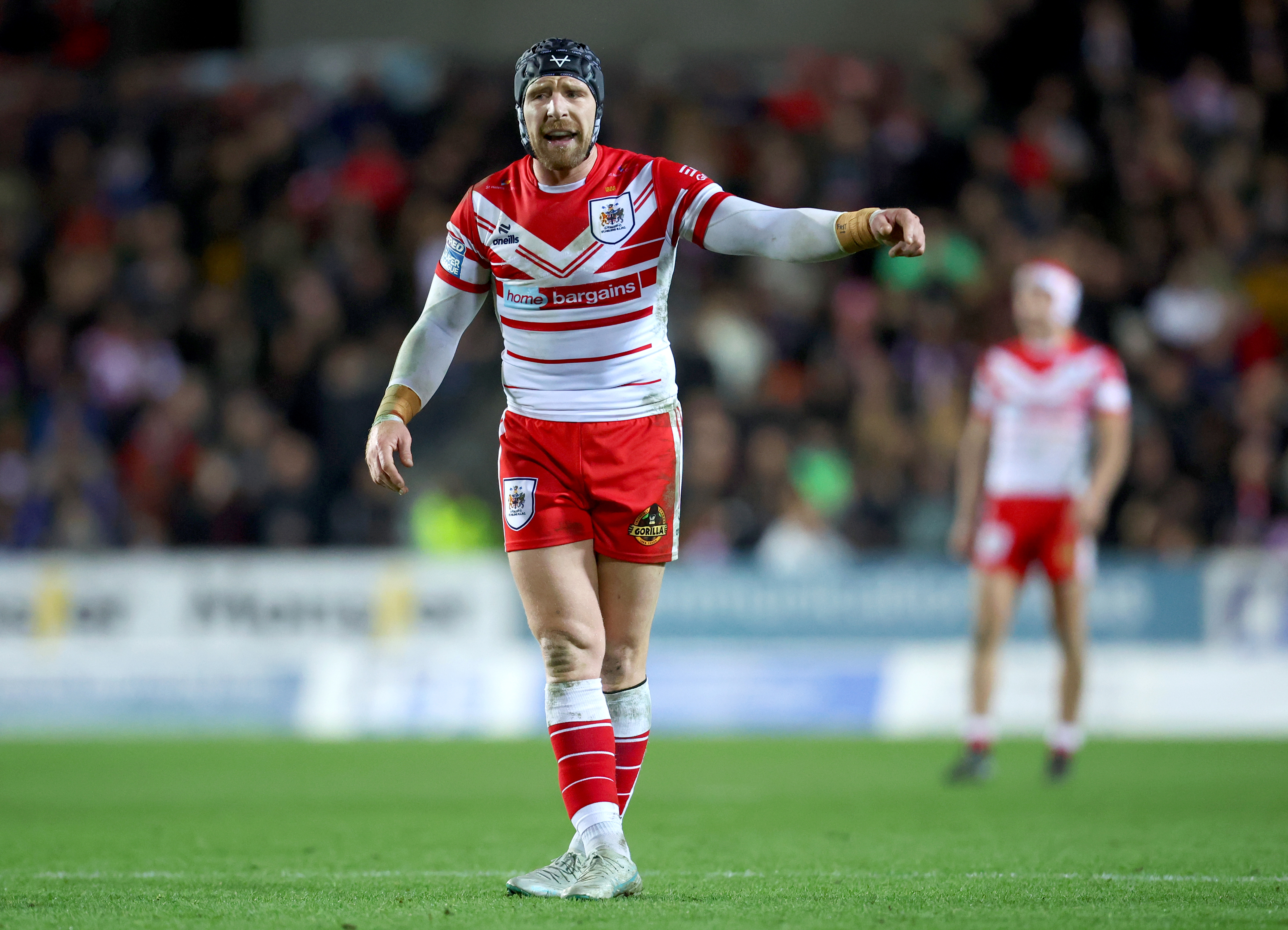 A rugby player for St Helens in a white and red jersey, black protective helmet, red shorts, and white socks with red stripes, standing on the field with his arm outstretched and an opponent out of focus behind him.
