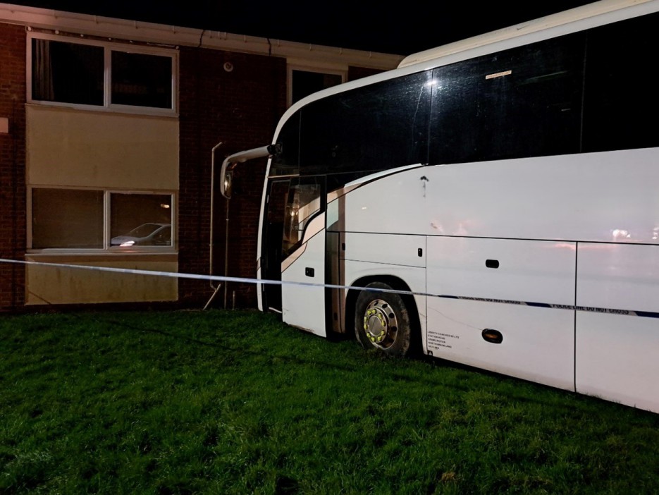 A large white coach bus with a damaged side is seen on a grassy area next to a brick building, cordoned off by "POLICE LINE DO NOT CROSS" tape.