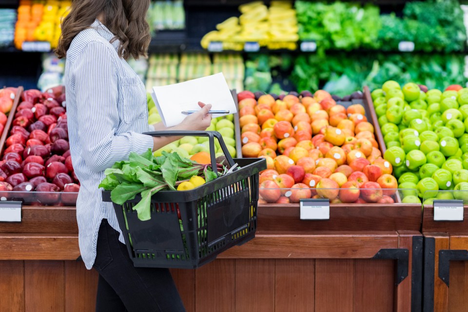Woman holding a grocery basket and a shopping list in a produce aisle.