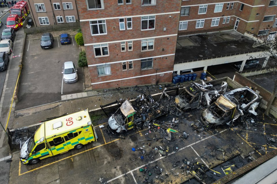 Aerial view of a parking lot showing several burned-out ambulances next to an apartment building, with one ambulance less damaged at the front left.