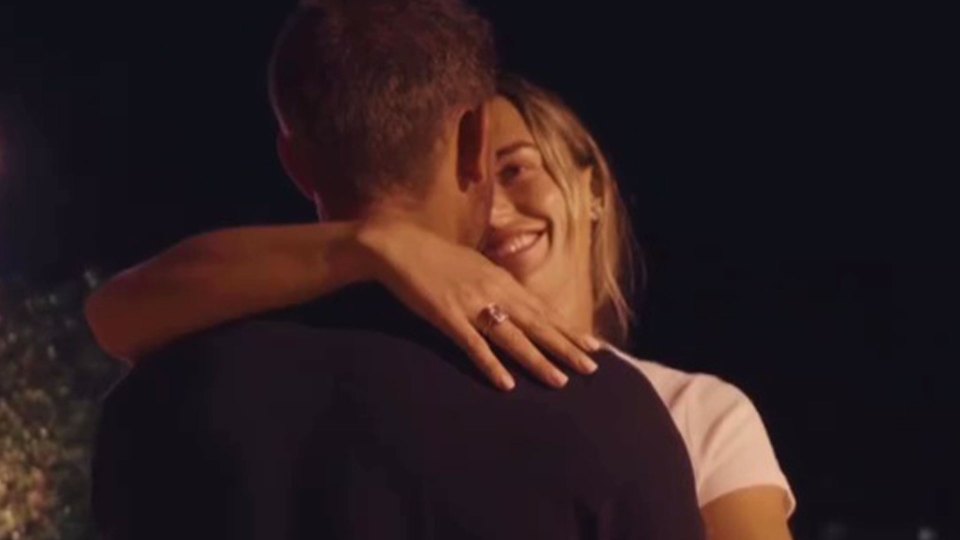 Woman with blonde hair smiling while hugging a man, wearing a large ring on her left ring finger.