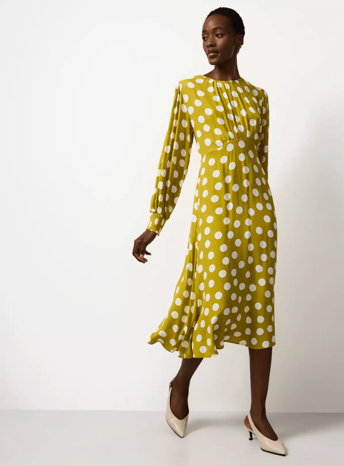 A dark-skinned woman in a flowing, long-sleeved chartreuse dress with white polka dots and white heels.