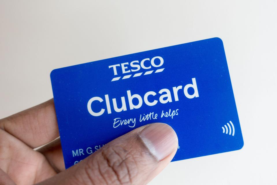 A person holding a blue Tesco Clubcard with the slogan "Every little helps" and a contactless payment symbol.