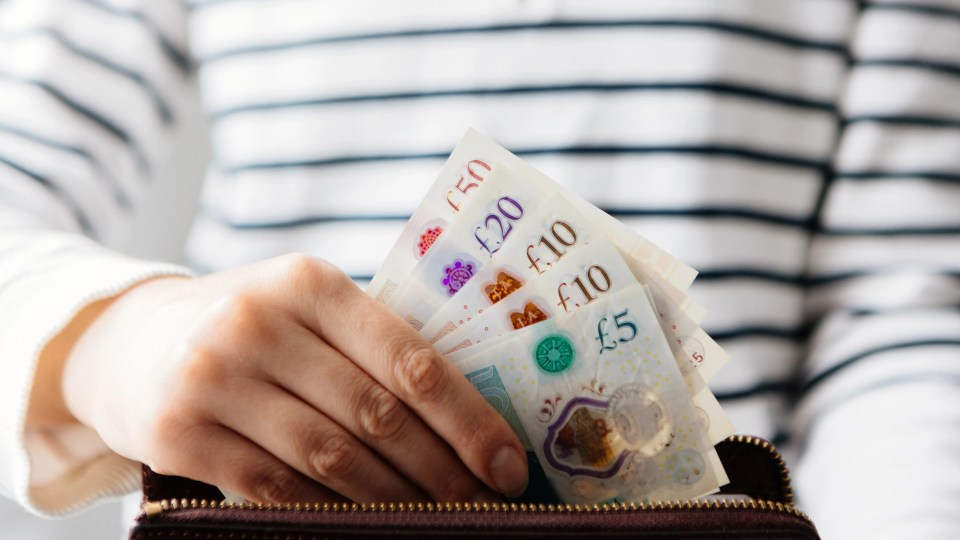 A person's hands pulling British currency notes (fivers, tenners, twenties, and fifties) from a wallet.