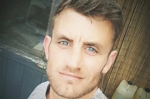 Paul Stewart Miller, a man with light brown hair, blue eyes, and a light beard, smiles faintly.