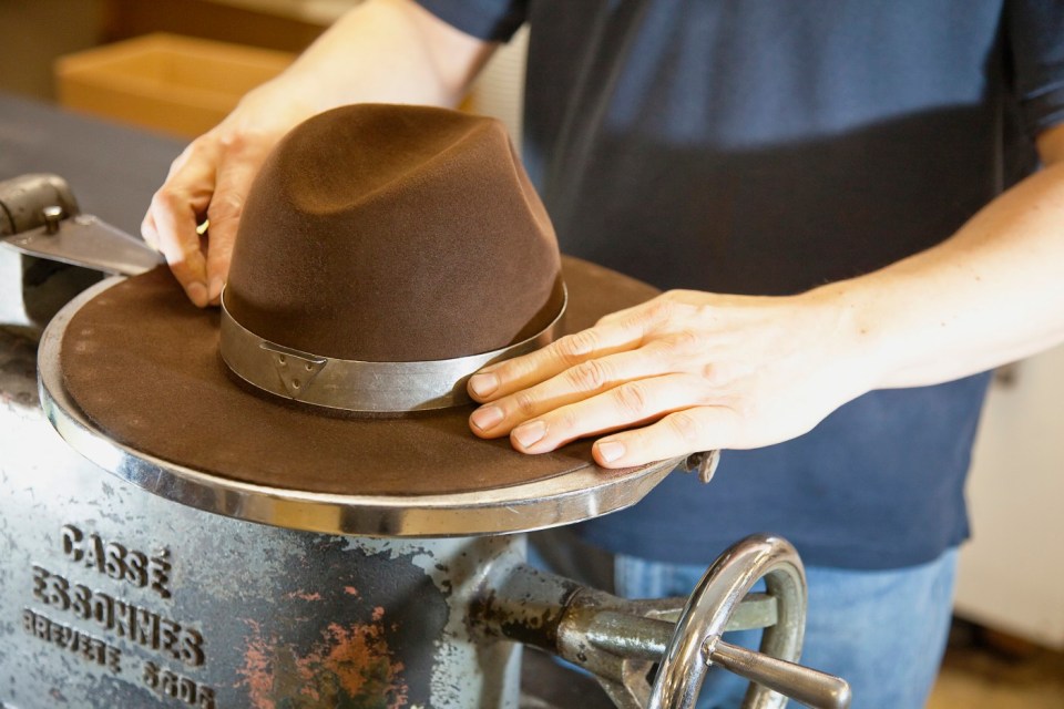 Hands shaping a brown felt hat on a specialized machine.
