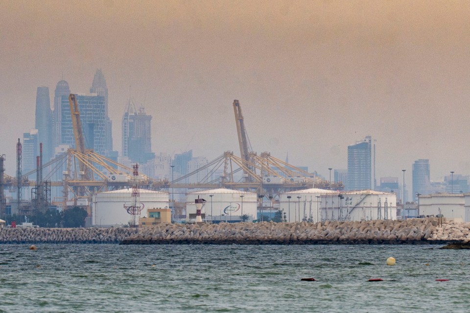 Oil tankers and cranes on the waterfront of Dubai's port, with a hazy skyline of tall buildings in the background.