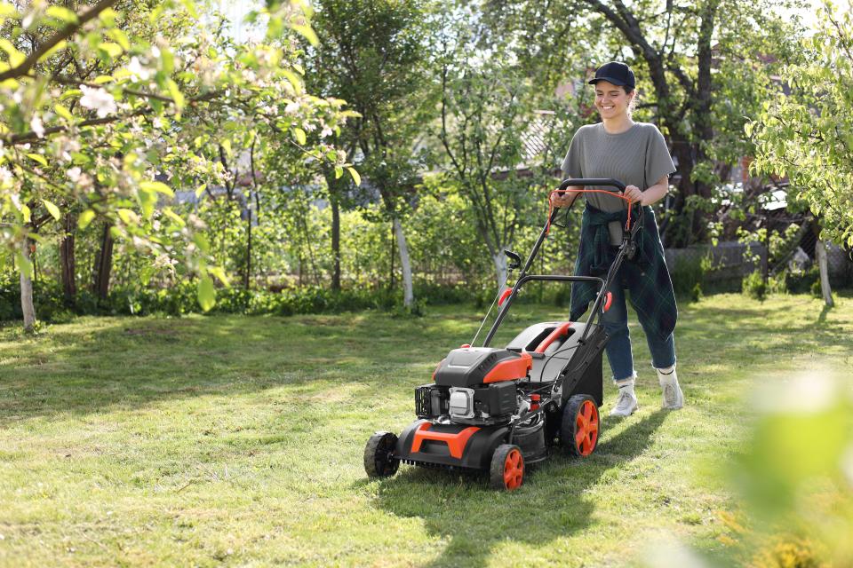 A smiling woman in a black baseball cap pushing a lawnmower across a green lawn.