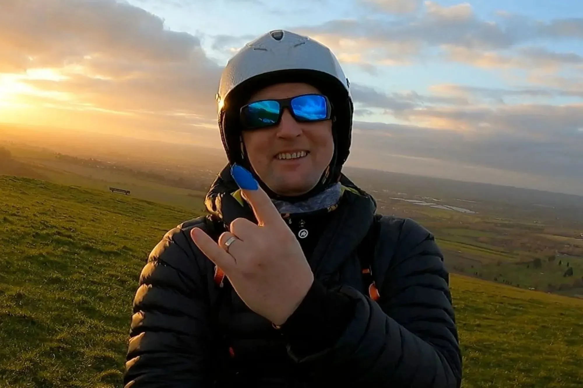 An image collage containing 1 images, Image 1 shows Man in helmet and sunglasses making a hand gesture with a bright sunset behind him over a green hillside