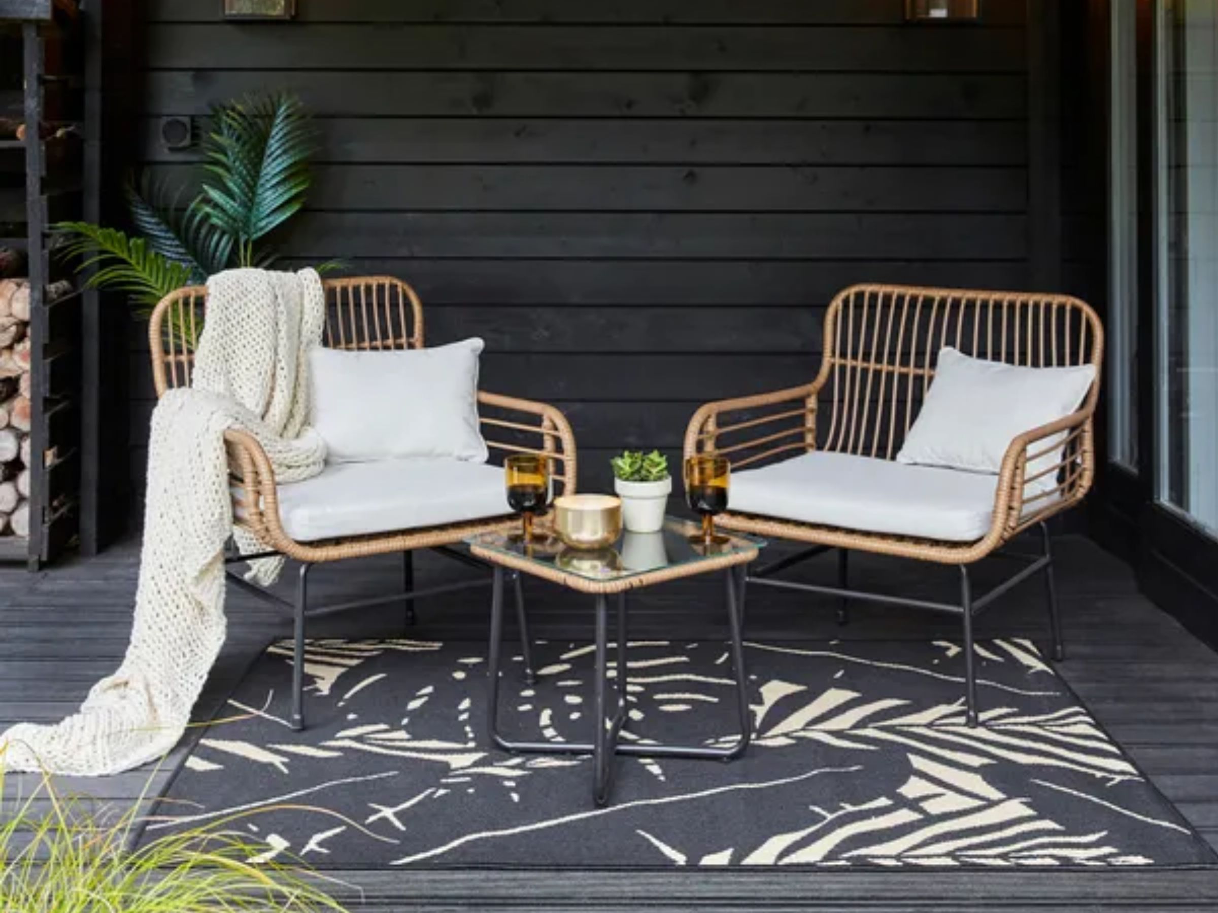 A patio with two wicker chairs, a glass-top table, and a black rug with white tropical leaves.