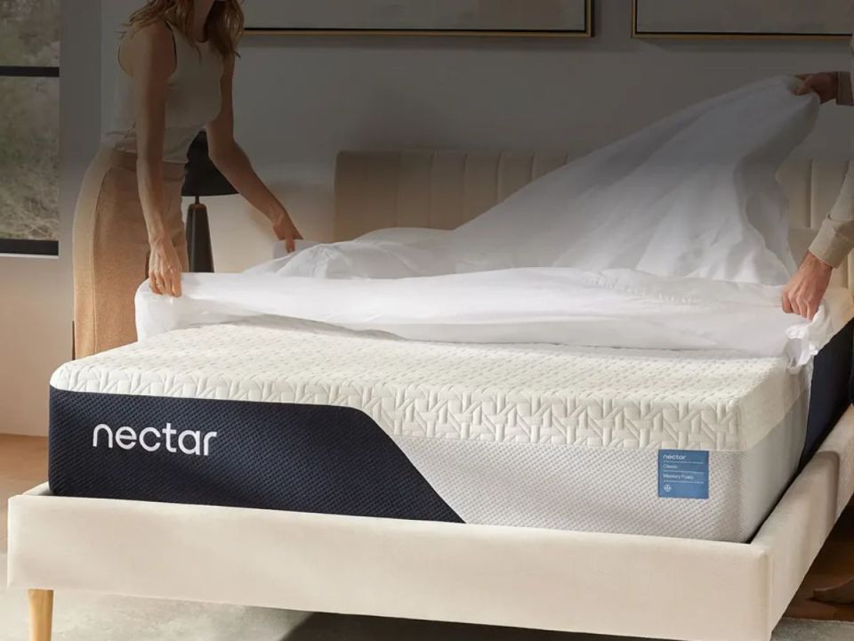 Two people putting a white sheet on a Nectar mattress in a bed frame.
