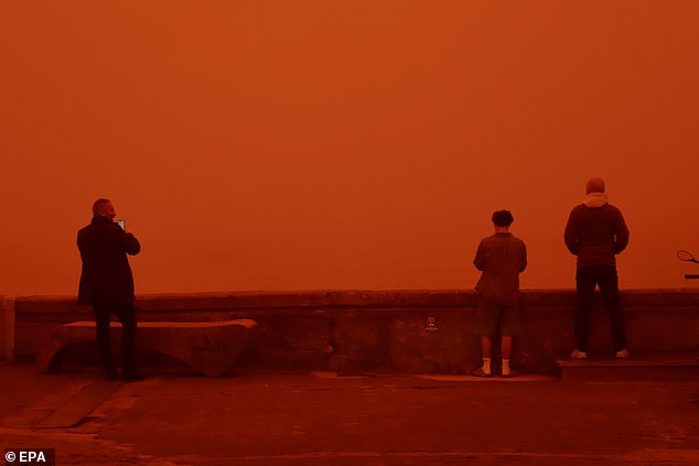 Three people look on from a lookout point as the red dust fills the sky