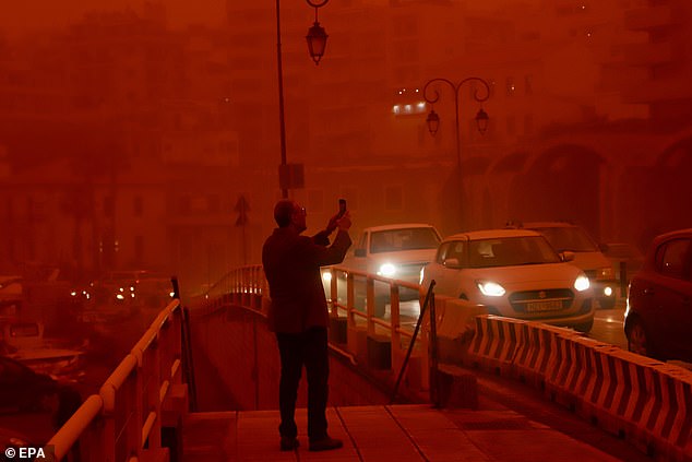 A man takes pictures of the surreal view of the city as drivers turn on their headlights to try and see through the dust