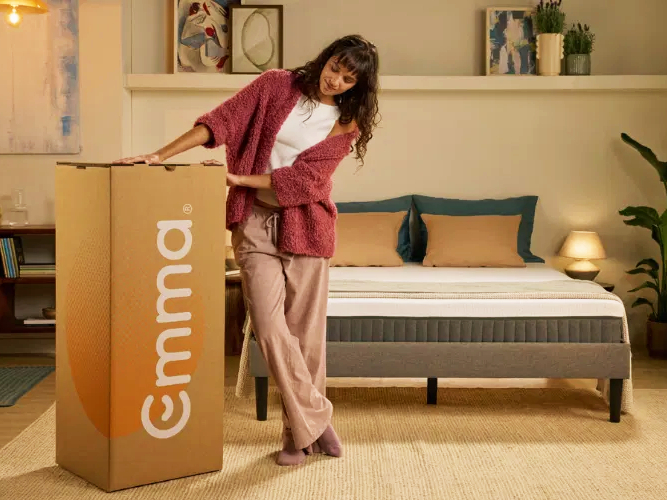 A woman in a living room next to a large Emma mattress box, with a new mattress visible on a bed behind her.