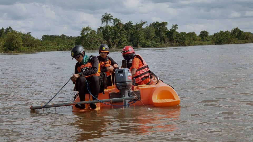 Three people in a small orange boat search a muddy river.