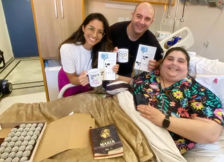 Three people, two visitors and one patient, smile while holding mugs and books in a hospital room.