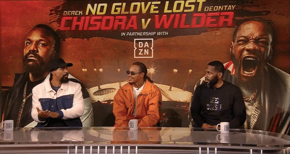Three men sit at a table in front of a banner advertising a "Chisora v Wilder" boxing match.