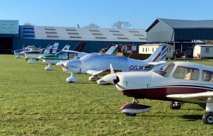 An image collage containing 1 images, Image 1 shows Small propeller planes parked on a grassy airfield