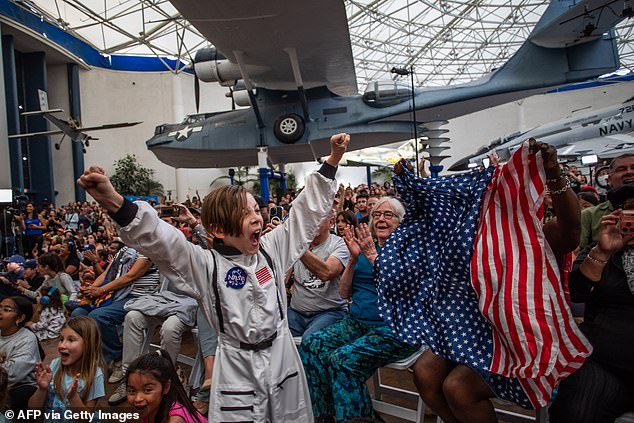 A young boy wearing an astronaut costume cheers next to a woman waving a flag as they watch a live broadcast of the return of the Artemis II crew members to Earth at the San Diego Air and Space Museum