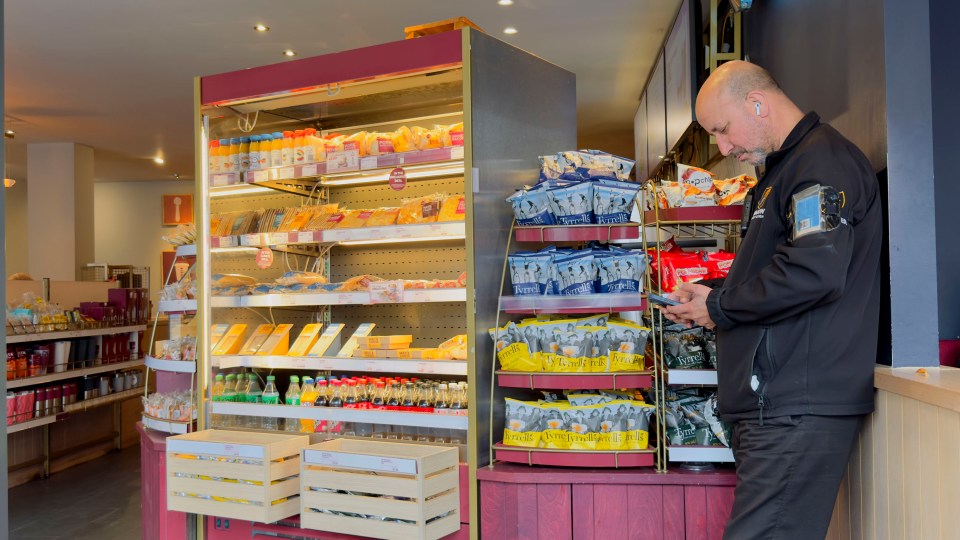 A security guard looking at his phone next to food and drink shelves in a Costa Coffee shop.