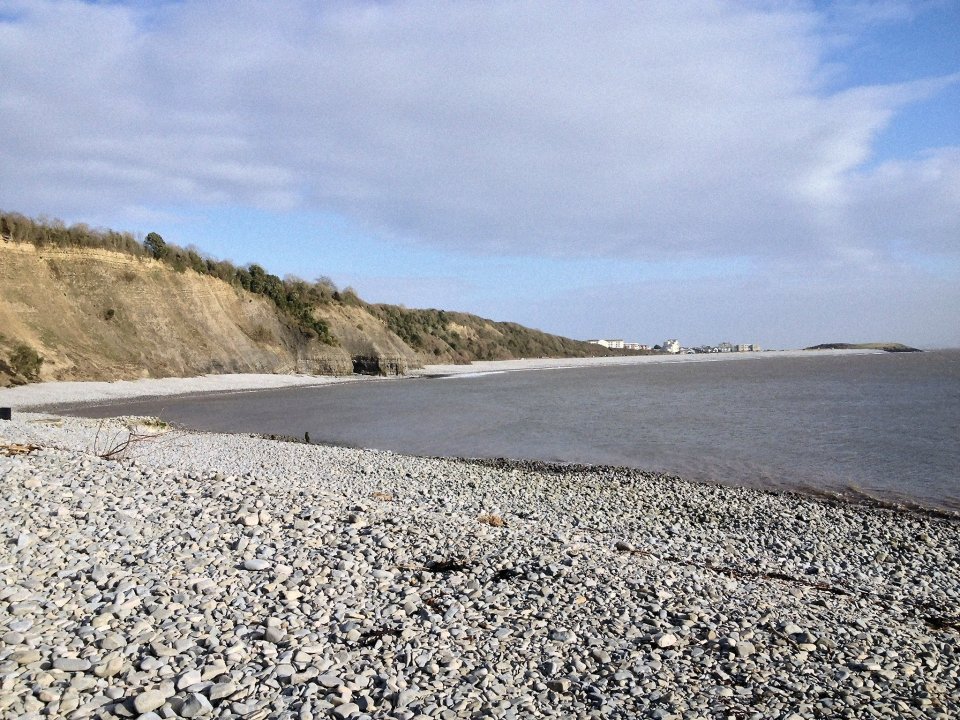 A wide view of a pebble beach next to the sea, with a rocky cliff covered in trees to the left, and distant buildings on the right horizon.
