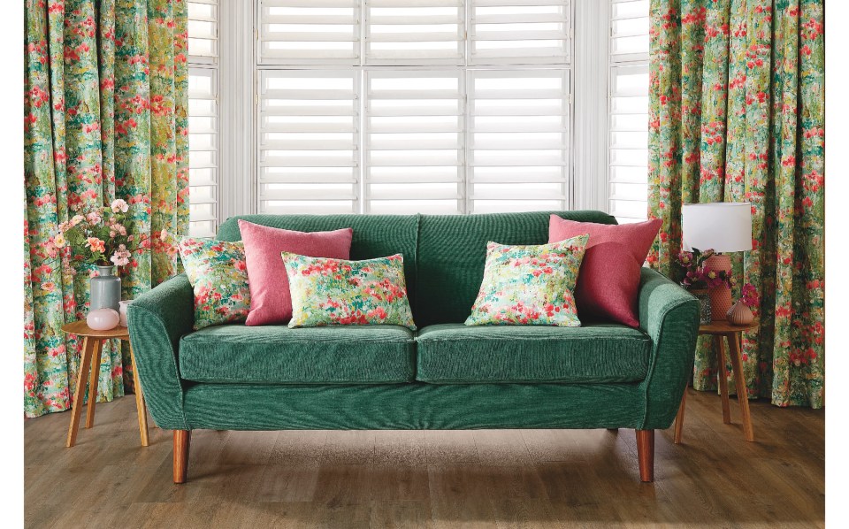 A green sofa with floral and pink cushions in a room with floral curtains and white wooden blinds.