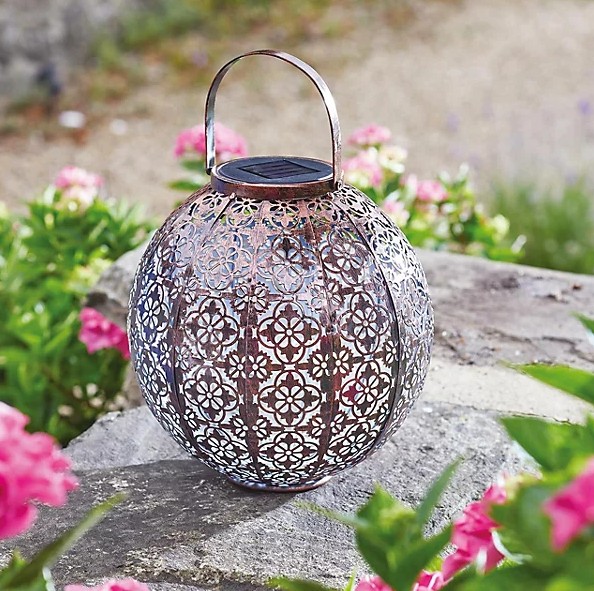 A round, bronze-colored solar-powered lantern with intricate cut-out patterns, sitting on a stone wall amidst pink flowers.